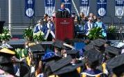 A man in academic regalia speaks to students before students wearing robes and mortarboards.